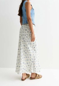 Denim mouwloze top gecombineerd met een vloeiende witte maxi rok met een blauw bloemmotief en bruine sandalen met twee banden en een kurken zool.