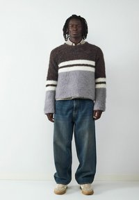 Flauschiger Strickpullover in Braun, Grau und Weiß mit Streifen; kombiniert mit lockeren blauen Denim-Jeans und beigen Turnschuhen.