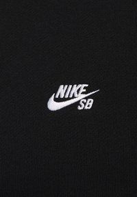 Tessuto nero con una texture a righe che presenta un logo Nike SB ricamato in bianco nell'angolo in alto a destra. Il design è semplice e minimalista.