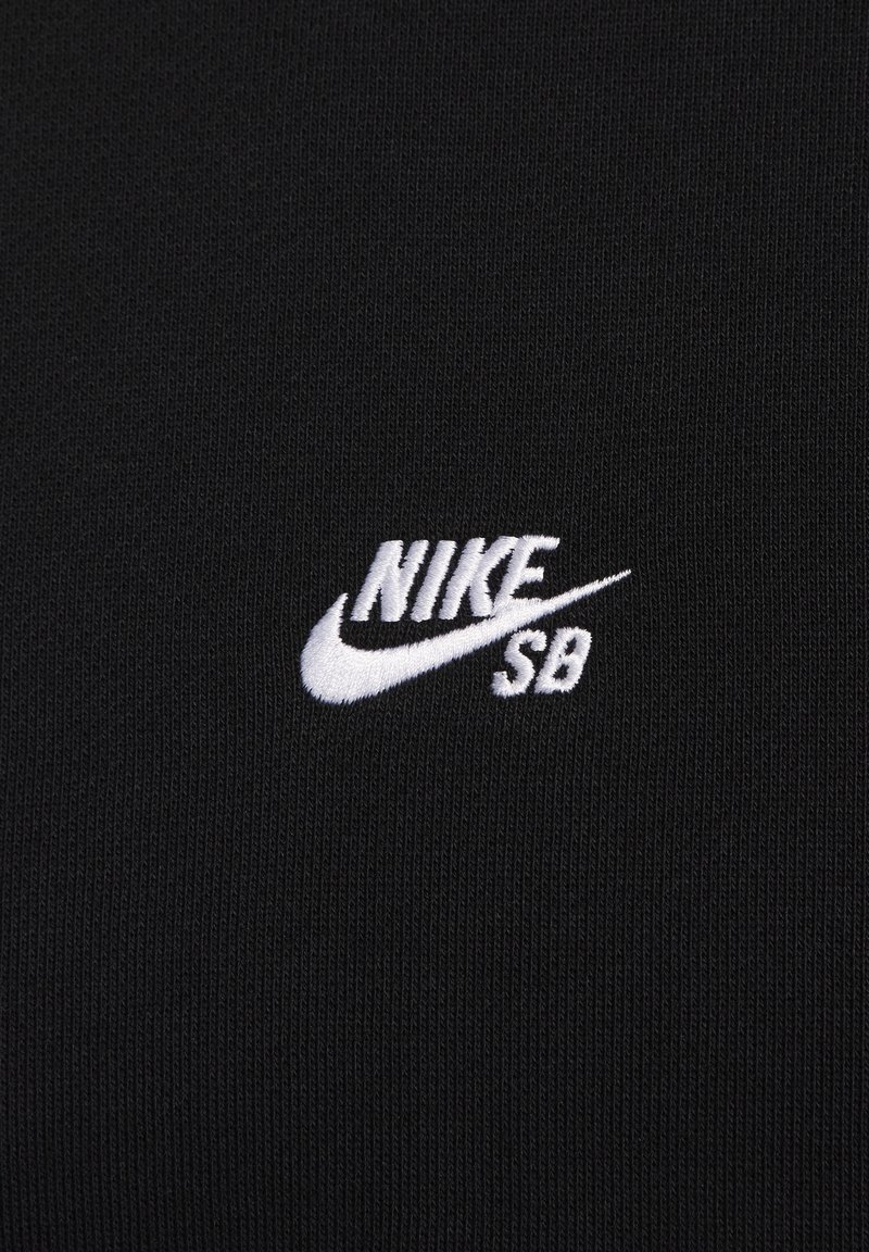 Svart tyg med ribbad textur, som har en broderad vit Nike SB-logotyp i övre högra hörnet. Designen är enkel och minimalistisk.
