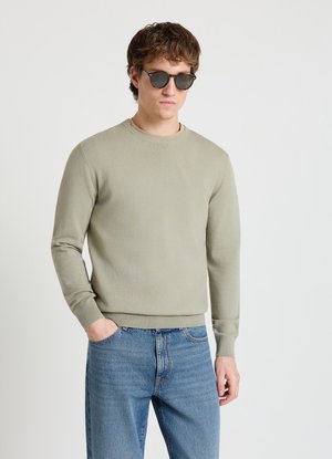 Giovane uomo con occhiali da sole scuri, maglione grigio chiaro girocollo e jeans blu, in piedi con una mano in tasca su sfondo semplice.