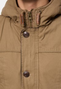 Brun huva jacka med dragkedja, två läderdetaljer och mörka knappar, med en slät, strukturerad tygfinish.