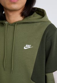 Közeli kép egy személyről, aki zöld Nike kapucnis pulcsit visel. A pulcsi fehér logóval és kontrasztos zöld textil szakaszokkal rendelkezik.