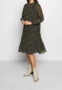 Robe noire à motifs floraux avec manches longues en tissu transparent, col montant et ourlet évasé. Portée avec des bottines brillantes et texturées.