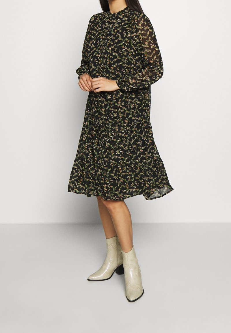 Robe noire à motifs floraux avec manches longues en tissu transparent, col montant et ourlet évasé. Portée avec des bottines brillantes et texturées.