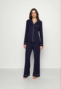 Kvinna som står barfota iförd en marinblå långärmad pyjamasoverall med vit piping och matchande lösa byxor mot en vit bakgrund.