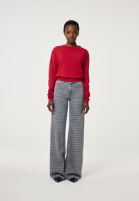 Eine Frau steht da und trägt ein rotes Langarmoberteil und weite graue gemusterte Hosen über schwarzen spitzen Schuhen vor einem schlichten weißen Hintergrund.