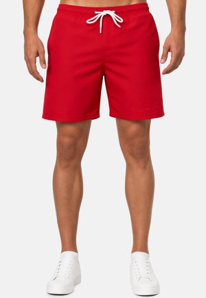 Homme portant un short de bain rouge vif avec un cordon blanc et des baskets blanches, debout devant un fond uni.