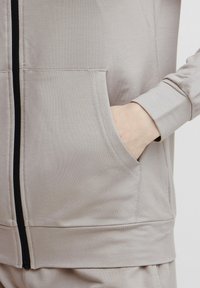 Svijetlosivi zip-up dukserica s prednjim džepom, rebrastim manžetama i teksturiranom tkaninom. Zipper se kontrastira s tamnim obrubom duž prednje strane.