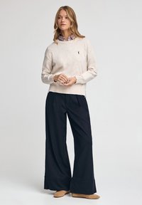 Beżowy sweter dziany z prążkowanymi mankietami, w połączeniu z szerokimi czarnymi spodniami i beżowymi butami wsuwanymi. Subtelne logo na swetrze.