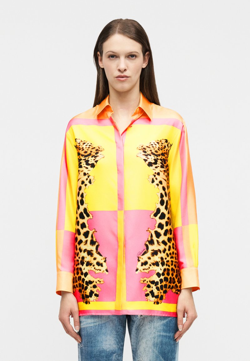 Γυναίκα μοντέλο με ίσια καστανά μαλλιά που φοράει πουκάμισο με κουμπιά, με κίτρινες, ροζ και λεοπάρ ύφασματικές λεπτομέρειες, συνδυασμένο με ξεθωριασμένο τζιν μπλε παντελόνι.