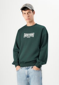 Man wearing a dark green sweatshirt with "HOMETOWN CHAMPION" text, light blue jeans, and a light gray baseball cap, hands in pockets.