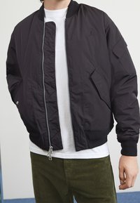 Svart bomberjacka i lätt material, med framtill dragkedja, ribbade ärmslut och nederkant, samt sidofickor. Logotyp på bröstet.