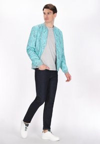 Giacca bomber azzurro chiaro con un motivo grafico, abbinata a una maglietta grigia, jeans scuri e sneakers bianche. Tessuto morbido, vestibilità rilassata.