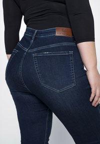 Donkerblauwe denim jeans met een gladde textuur; heeft achterzakken en een leren merklabel in bruin aan de taille.