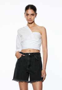 Vit omslagsskjorta med en lång ärm, parat med högmidjade svarta denimshorts med fransade kanter. Modellen står med händerna i fickorna.
