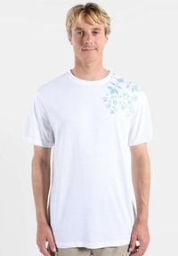 T-shirt en coton blanc avec un motif floral bleu clair et bleu sarcelle sur une épaule. Manches courtes, encolure ronde, coupe décontractée.