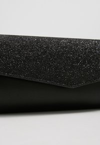 Sac à main noir avec un panneau supérieur pailleté noir et une section inférieure lisse noire, présentant un design de rabat angulaire et net.