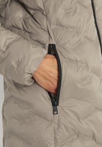 Pufferjack in lichtgrijze stof met een gewatteerde textuur. Heeft een zwarte ritszak aan de zijkant, waar een hand in reikt.