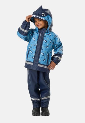 Enfant souriant en imperméable et pantalon bleus sur le thème du requin, tenant la capuche avec un visage de requin, debout devant un fond uni.
