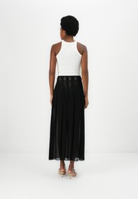 Gonna maxi nera plissettata con pannelli trasparenti, abbinata a un top aderente bianco. Vista da dietro, mostra il design e il movimento.