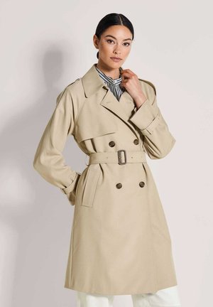 Frau mit dunklen Haaren trägt einen beigen trenchcoat mit Gürtel und ein gestreiftes Hemd, posiert mit einer Hand in der Nähe des Kragens vor einem einfarbigen Hintergrund.