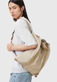 Mochila de tela beige con secciones acolchadas, correas en contraste de color verde oliva y detalles de costura roja. Lleva puesta sobre un hombro.