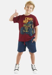 Bordeaux katoenen t-shirt met een kleurrijke monstertruckafbeelding, gecombineerd met marineblauwe shorts en zwarte hoge sneakers.