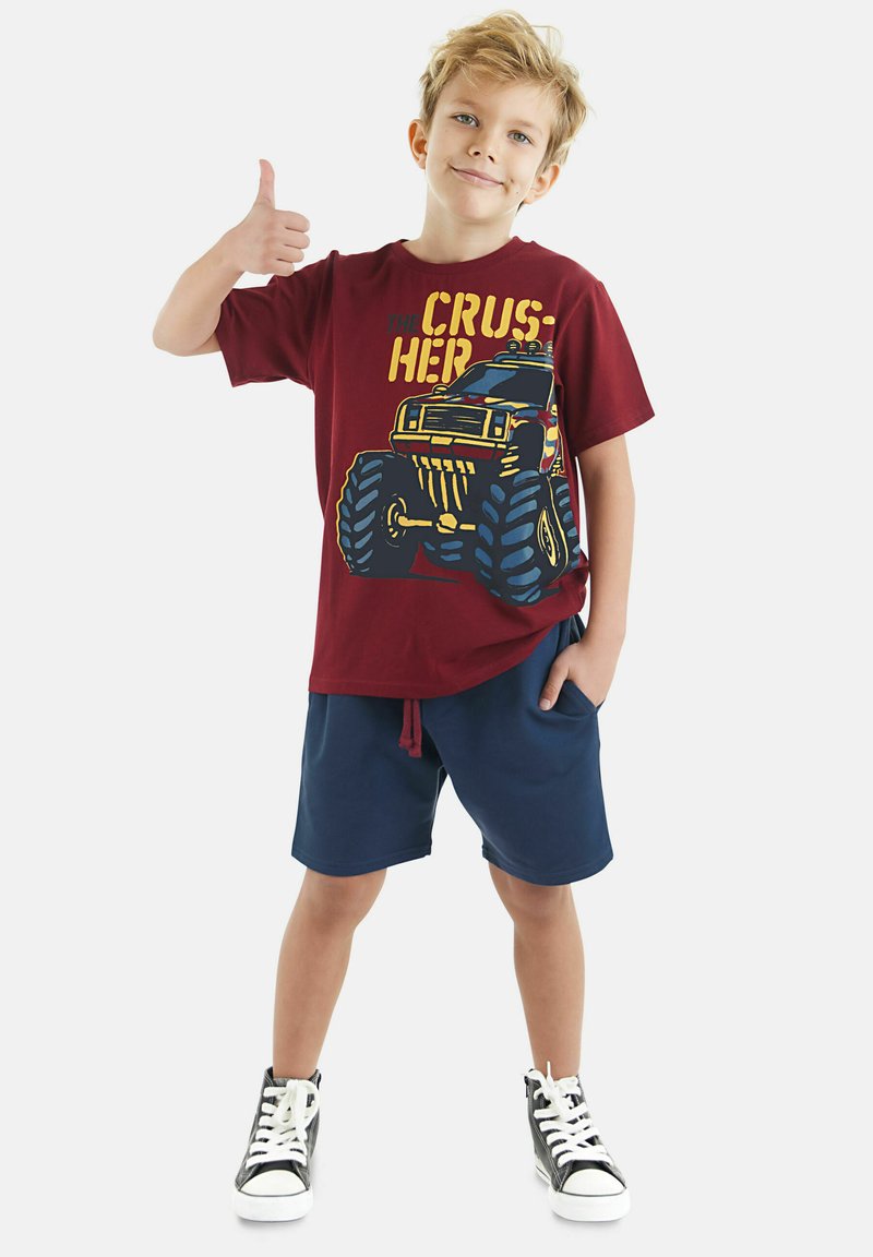 Bordeaux katoenen t-shirt met een kleurrijke monstertruckafbeelding, gecombineerd met marineblauwe shorts en zwarte hoge sneakers.