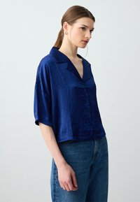Satinbluse in Marineblau mit kurzen Ärmeln, geformtem Kragen, Knopfleiste und leicht strukturiertem Stoff. Kombiniert mit hochgeschnittenen Jeans.