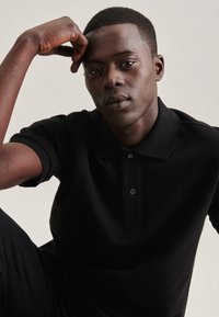 Zwarte polo met een klassieke kraag, drie knopen en korte mouwen, gedragen door een zittend persoon met kort haar.