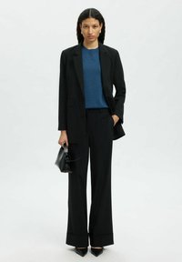 Musta räätälöity bleiseri, joka on yhdistetty siniseen neulepaitaan, mustiin leveälahkeisiin housuihin, teräväkärkisiin mustiin korkokenkiin ja pieneen mustaan käsilaukkuun.