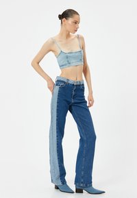 Top in denim blu chiaro, corto, abbinato a jeans a vita alta con un pannello laterale blu chiaro. La modella è in piedi con una mano sulla vita.