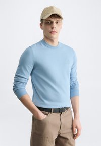 Jeune homme portant une casquette beige, un pull bleu clair et un pantalon kaki avec une ceinture noire, debout avec une main dans la poche sur fond blanc.