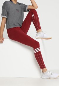 Persona sentada en un bloque blanco, vistiendo leggings color burdeos con rayas blancas y zapatillas blancas, combinados con una camiseta gris de manga corta.