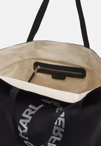 Svart tote-väska med en krämfärgad insida. Har en dragkedjeficka och en reflektiv logotryck på yttertyget.