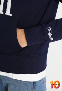 Marineblauwe sweatshirt met witte trekkoorden, voork pocket en geborduurde "Original" details op de mouw; zachte katoenen stofmix.