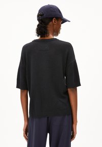 Schwarzer gerippter Pullover mit kurzen Ärmeln, lockerer Passform und einem kleinen Etikett auf der Rückseite. Kombiniert mit einer dunkelblauen Hose und einer passenden Mütze.