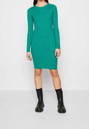 Robe à manches longues côtelée vert sarcelle, silhouette ajustée, encolure ronde, tissu en maille texturée, portée avec des bottines noires.