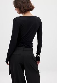 Schwarzes Langarmoberteil mit rundem Ausschnitt, das eine glatte Textur aufweist. Getragen mit maßgeschneiderten schwarzen Hosen und silbernen Armreif-Akzenten.