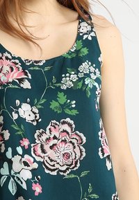 Débardeur floral en tissu bleu-vert présentant de grandes roses roses et blanches, des feuilles vertes et de petites fleurs blanches. Encolure dégagée et sans manches.