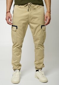 Pantalons cargo beiges avec des poignets élastiques, poches latérales et accents noirs. Fabriqués en tissu léger, dotés d'une taille à cordon de serrage pour un ajustement réglable.