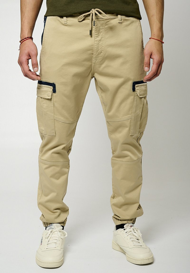 Pantalons cargo beiges avec des poignets élastiques, poches latérales et accents noirs. Fabriqués en tissu léger, dotés d'une taille à cordon de serrage pour un ajustement réglable.