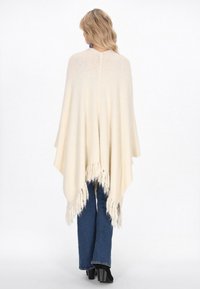 Poncho de culoare crem, cu un tiv franjurat, realizat dintr-un material moale, având un design drapat care se extinde peste umeri și spate.