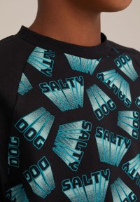 Čierne tričko s opakujúcim sa tyrkysovým grafickým vzorom so slovami "SALTY" a "DOG", navrhnutými v rôznych orientáciách.