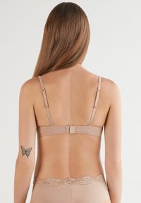 Beżowy biustonosz z regulowanymi ramiączkami i zapięciem na haftkę. Teksturowany materiał z koronkowym wykończeniem wzdłuż dolnej krawędzi. Prosty i funkcjonalny design.