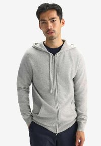 Ljusgrå zip-up hoodie gjord av mjukt tyg, med en framsida dragkedja, huva och sidofickor. Den har ribbstickade muddar och fåll.
