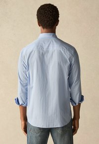 Cămaș�ă albastru deschis cu mâneci lungi, cu dungi verticale albe, manșete cu nasturi răsucite, și tiv curbat. Material moale pe un fundal neutru.