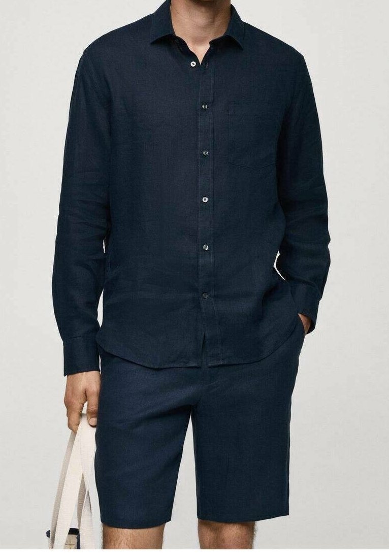 Homme portant une chemise bleu marine foncé à manches longues boutonnée et un short assorti jusqu'aux genoux, tenant un sac fourre-tout beige dans une main.