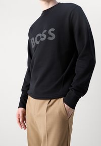 Felpa nera con grande logo grigio "BOSS" sulla parte anteriore. Vestibilità regolare con collo tondo e polsini a costine. Abbinata a pantaloni beige.